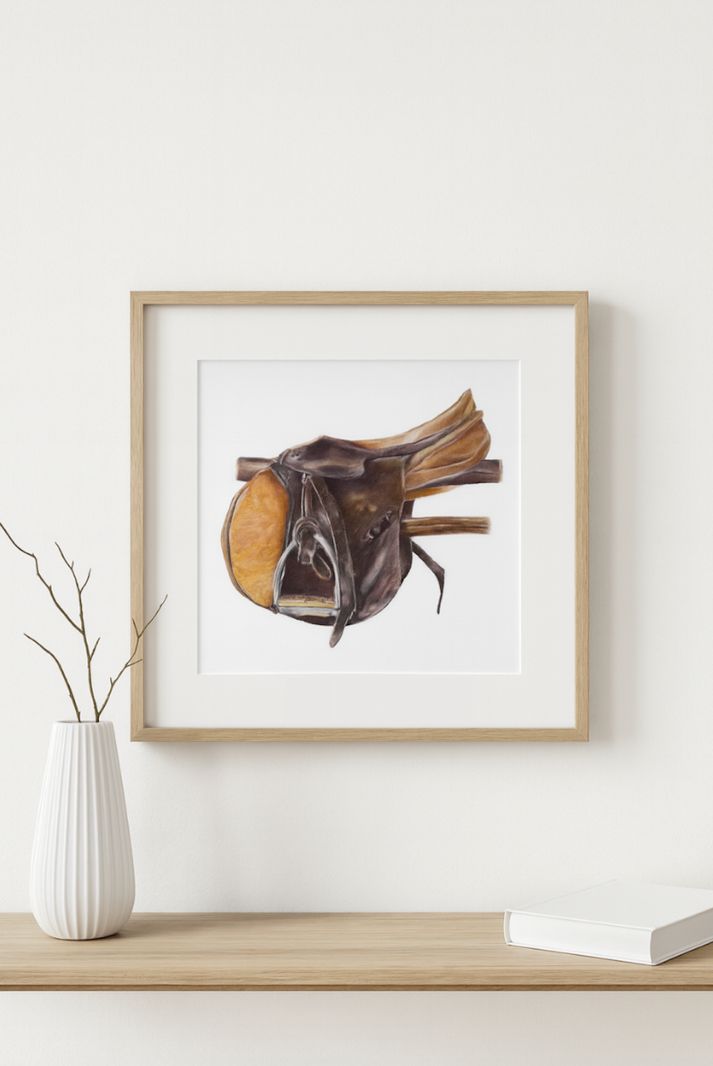 Framed artwork of a horse saddle on a white wall with a vase and books below.